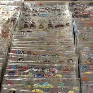May include: A display of various sticker sheets in clear plastic packaging. The stickers feature a variety of designs, including flowers, cartoon characters, and text. The stickers are in different colors and sizes, and are arranged in rows.
