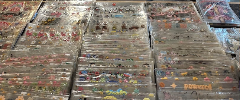 May include: A display of various sticker sheets in clear plastic packaging. The stickers feature a variety of designs, including flowers, cartoon characters, and text. The stickers are in different colors and sizes, and are arranged in rows.