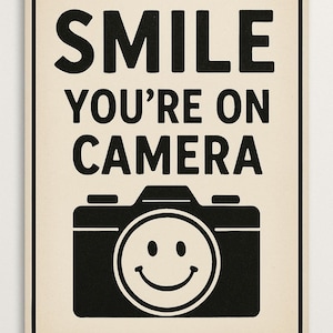 May include: A rectangular sign with a cream background and black text and graphics. The text reads "SMILE YOU'RE ON CAMERA" above a camera graphic with a smiley face in the lens.