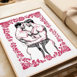 May include: A framed art print featuring a black and white ink-style illustration of two muscular figures embracing, surrounded by a decorative border of vibrant pink roses. The artwork is presented in a light-colored wooden frame on a flat surface.