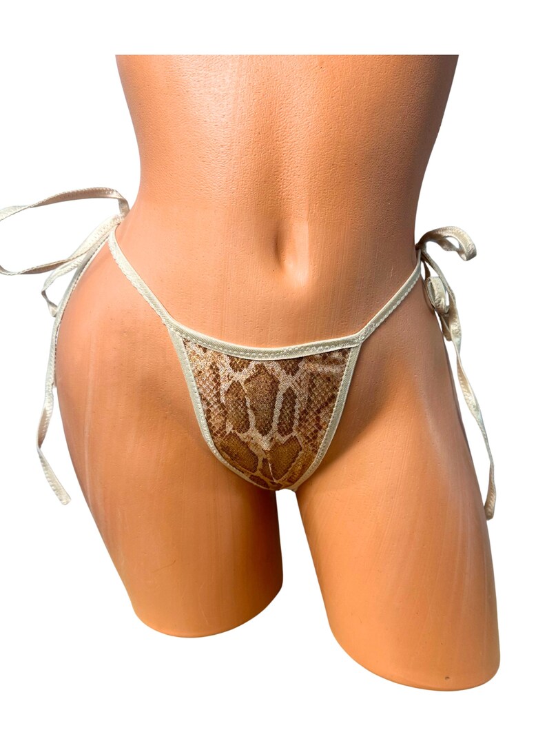 K&ouml;nnte beinhalten: Ein Bikiniunterteil mit einem braun-beigen Schlangenledermuster. Das Kleidungsst&uuml;ck verf&uuml;gt &uuml;ber d&uuml;nne, helle seitliche Bindeb&auml;nder zur Anpassung und ein dreieckiges Vorderteil mit passender heller Einfassung an den Stoffkanten.