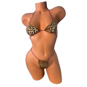 May include: A two-piece bikini set with a leopard print pattern. The top is a triangle style with thin red straps. The bottom is a thong style, also with the leopard print. The bikini is displayed on a mannequin.