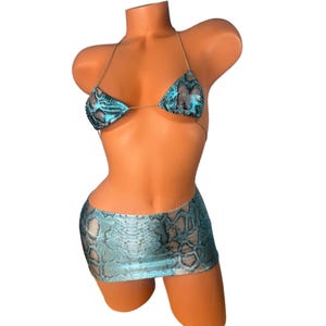 Puede incluir: Un conjunto de bikini con estampado de piel de serpiente turquesa, que incluye un top triangular y una minifalda. El top del bikini tiene finas correas doradas. El conjunto se muestra en un maniquí.
