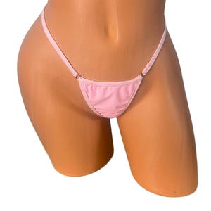 May include: A pale pink thong bikini bottom. The design features a triangular front and delicate straps that attach to the sides with small metal rings. The fabric appears smooth, and the overall style is minimalist.