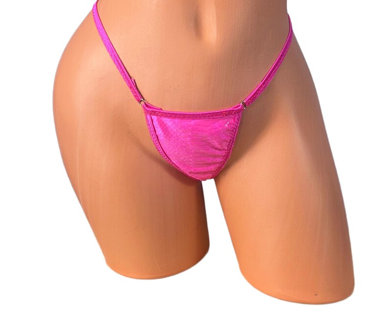 K&ouml;nnte beinhalten: Ein leuchtend pinkfarbener String-Bikini-Slip. Der Stoff hat ein strukturiertes Aussehen. Der Bikini-Slip wird auf einer Schaufensterpuppe pr&auml;sentiert. Die Tr&auml;ger sind d&uuml;nn und zart.