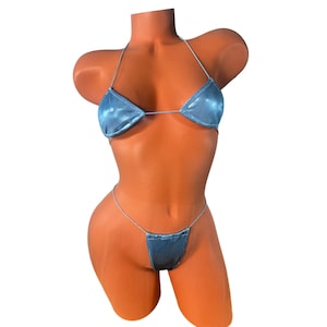 May include: A light blue bikini set on a mannequin. The top is a triangle style with thin straps, and the bottom is a matching thong. The fabric has a slight sheen. The bikini is designed for swimwear.