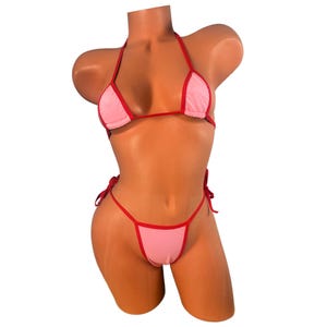 May include: A two-piece bikini set in light pink with red trim. The triangle top and string bottom are displayed on a mannequin. The bikini features tie closures on the top and bottom. The overall style is minimalist and designed for swimwear.
