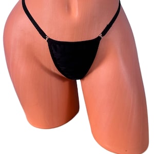 Puede incluir: Tanga negro con tirantes finos y pequeños anillos metálicos en los laterales. La ropa interior está hecha de una tela brillante y suave. La prenda se exhibe en un maniquí.