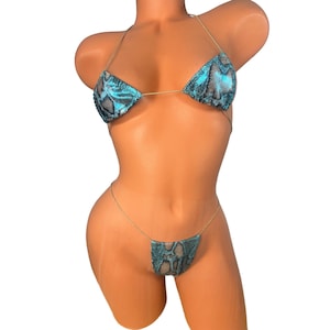 Può includere: Un set bikini con stampa a pelle di serpente turchese e nero. Il top a triangolo e lo slip a perizoma sono collegati da sottili spalline color oro. Il costume da bagno è esposto su un manichino.