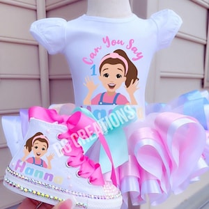 Peut inclure: Ensemble pour tout-petit blanc avec un tutu et des baskets assortis. Le haut présente un dessin animé et le texte "Can You Say 1". Les baskets sont ornées de strass et d'un nœud rose. Le tutu est rose, blanc et bleu.
