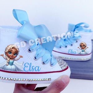 May include: White high-top sneakers featuring a cartoon image of Elsa, the character from Frozen, with the name "Elsa" in blue script. The shoes have light blue ribbon laces and are embellished with rhinestones.