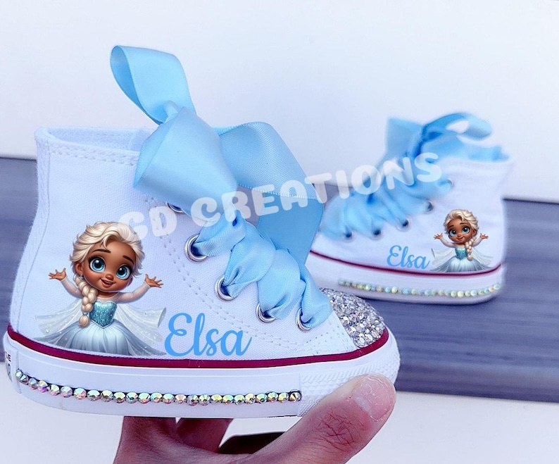 May include: White high-top sneakers featuring a cartoon image of Elsa, the character from Frozen, with the name "Elsa" in blue script. The shoes have light blue ribbon laces and are embellished with rhinestones.