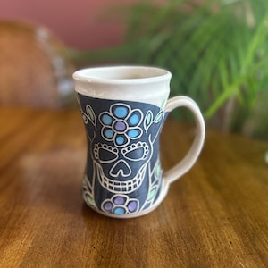 May include: A ceramic mug with a white handle and rim. The mug features a black background with a sugar skull design in white, blue, and purple. Floral accents in green and purple surround the skull. The mug sits on a wooden surface.