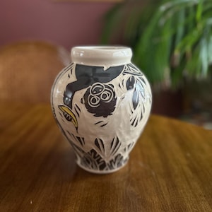 May include: A ceramic vase with a cream-colored base and a black and white jungle-themed design. The vase has a wide body and a narrow neck, with a white rim. The design includes monkeys, leaves, and bananas. The vase is sitting on a wooden table.