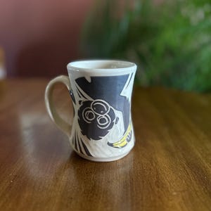 May include: A ceramic mug with a whimsical monkey design. The mug features a white base with a dark blue band around the top, adorned with a black and white monkey illustration and a yellow banana. The handle is a light beige color.
