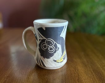 May include: A ceramic mug with a whimsical monkey design. The mug features a white base with a dark blue band around the top, adorned with a black and white monkey illustration and a yellow banana. The handle is a light beige color.