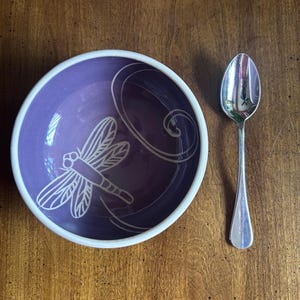 May include: A purple ceramic bowl with a white dragonfly design and a white rim. A silver spoon is placed to the right of the bowl. The bowl and spoon are on a wooden surface.