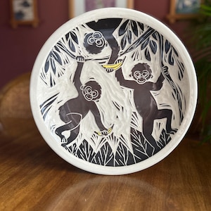 May include: A white ceramic bowl with a black and white design of three monkeys in a tree. The monkeys are holding bananas and are surrounded by leaves.