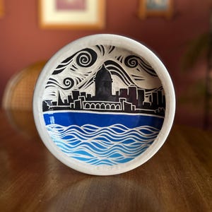 May include: White ceramic bowl with a blue and white design of a city skyline and wavy water. The skyline is black and the water is blue with white waves.