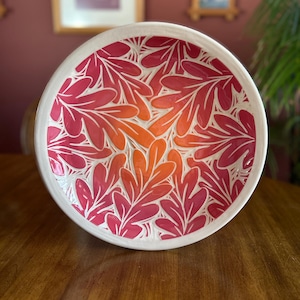 May include: A white ceramic bowl with a pink and orange leaf pattern. The bowl is decorated with a gradient of color, starting with a deep pink at the top and fading to a light orange at the bottom.