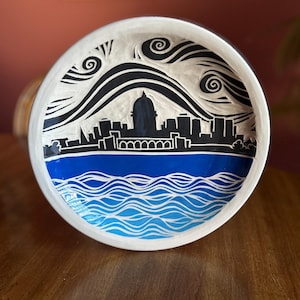 May include: A white ceramic bowl with a black and white cityscape design and blue waves at the bottom. The cityscape features a dome-shaped building in the center.