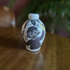 May include: A small, white and brown ceramic vase with a monkey design. The vase features a brown monkey holding a yellow banana, with brown leaves and a white background. The vase is on a wooden surface.