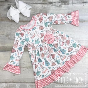 May include: A white long-sleeved dress with a pattern of dresses in pink, blue, and peach. The dress has pink gingham trim on the sleeves and hem, and a pink flower applique. A large white bow is placed near the dress.