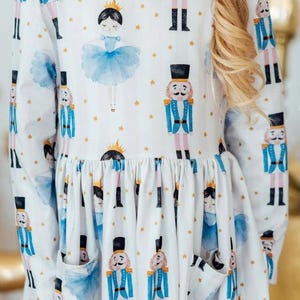 May include: White long-sleeved dress featuring a repeating pattern of nutcrackers, ballerinas, and gold stars. The dress has a gathered waist and two pockets. The nutcrackers are in blue and gold uniforms, and the ballerinas wear blue tutus.