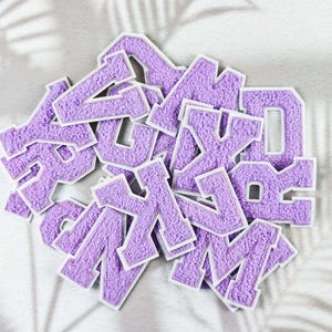 May include: A collection of purple chenille letter patches with white borders. The letters are in a varsity style and are scattered across a white surface. The image is well-lit, showcasing the texture and color of the patches.
