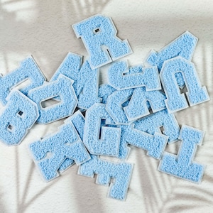May include: A collection of light blue, fuzzy fabric letters with white borders. The letters are scattered on a white surface, with shadows from a leafy plant in the background. The letters are in various fonts and sizes.