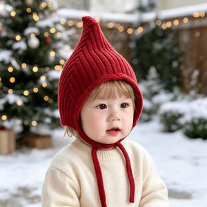 May include: A bright red knitted bonnet with a pointed top and a ribbed texture. The bonnet features two long red knitted ties that hang down from the chin area. The hat is worn over a cream-coloured long-sleeved sweater with a ribbed neckline.