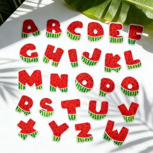May include: A complete alphabet of watermelon-themed letter stickers. Each letter is red with black seeds, a green rind, and a white border. The letters are arranged on a white surface with a banana leaf in the background.
