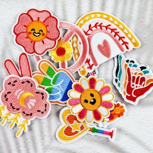 Könnte beinhalten: Eine Sammlung von gestickten Aufnähern mit Retro-Thema. Designs umfassen ein Friedenszeichen, einen Regenbogen, Blumen und eine Handgeste. Die Aufnäher haben leuchtende Farben wie Rosa, Gelb, Orange und Blau mit weißen Rändern.