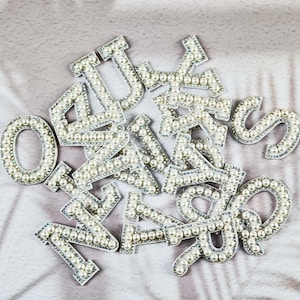 Könnte beinhalten: Eine Ansammlung silberfarbener Buchstaben- und Zahlenverzierungen, jede mit kleinen, weißen Kunstperlen besetzt. Die Buchstaben und Zahlen sind auf einer strukturierten, hellgrauen Oberfläche verstreut und bilden ein dekoratives Element.