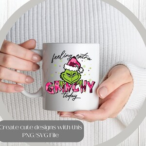 May include: White ceramic mug featuring a green Grinch character wearing a pink and white plaid Santa hat. The mug has the words "feeling extra Grinchy today" in black and pink. The mug is held by a person.