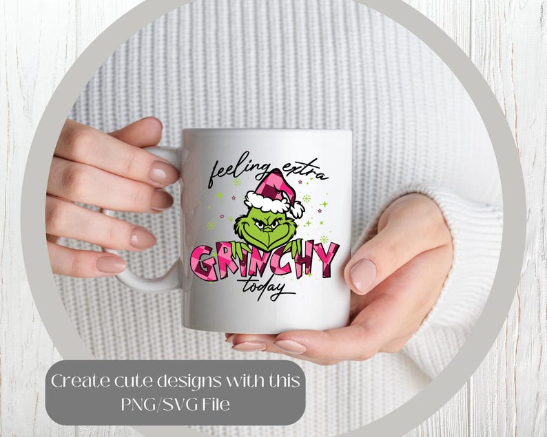 May include: White ceramic mug featuring a green Grinch character wearing a pink and white plaid Santa hat. The mug has the words "feeling extra Grinchy today" in black and pink. The mug is held by a person.
