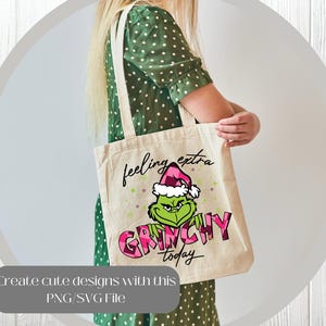 May include: A beige tote bag featuring a Grinch graphic wearing a Santa hat, with the words "feeling extra GRINCHY today." The Grinch is green, and the text is pink and black. The bag has a simple design with two handles.