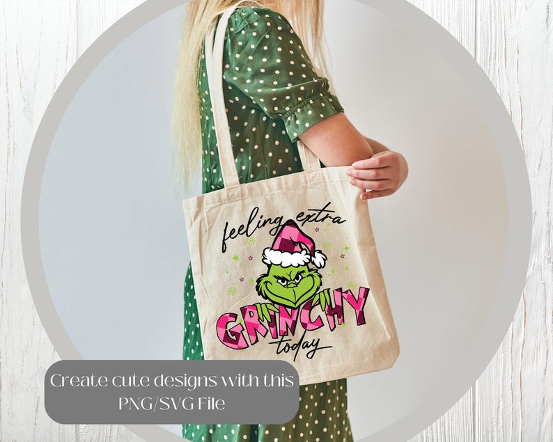 May include: A beige tote bag featuring a Grinch graphic wearing a Santa hat, with the words "feeling extra GRINCHY today." The Grinch is green, and the text is pink and black. The bag has a simple design with two handles.
