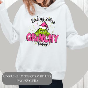 May include: White hoodie featuring a Grinch graphic wearing a Santa hat, with the text "feeling extra GRINCHY today" in pink and green. The design includes Christmas lights and stars. A PNG/SVG file is available.
