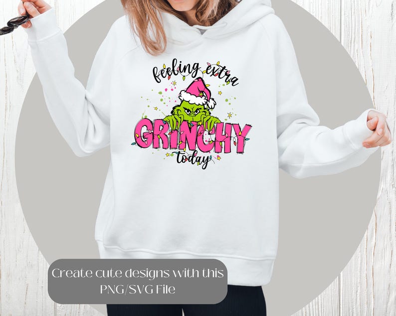 May include: White hoodie featuring a Grinch graphic wearing a Santa hat, with the text "feeling extra GRINCHY today" in pink and green. The design includes Christmas lights and stars. A PNG/SVG file is available.