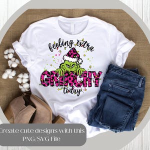 May include: White t-shirt with a Grinch graphic. The Grinch is green and wearing a pink and black leopard print hat. The words "feeling extra GRINCHY today" are printed on the shirt in black and pink.