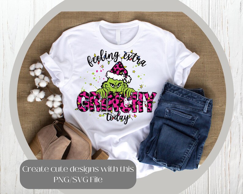 May include: White t-shirt with a Grinch graphic. The Grinch is green and wearing a pink and black leopard print hat. The words "feeling extra GRINCHY today" are printed on the shirt in black and pink.