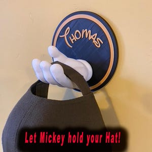 May include: A white Mickey Mouse hand wall hook with a black and orange circle behind it. The circle has the name "Thomas" written on it. A brown baseball cap is hanging from the hook. The text "Let Mickey hold your hat!" is written on the cap.