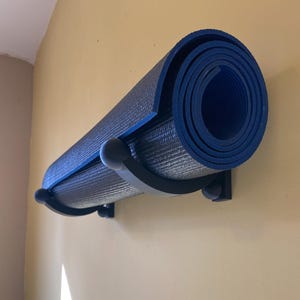 May include: A rolled-up blue yoga mat is held by a black metal wall mount. The mat is secured by two curved brackets and small ball-shaped ends. The wall is painted a light yellow color.