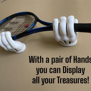 Pode incluir: Uma raquete de tênis segurada por um par de mãos brancas de desenho animado. A raquete tem uma estrutura preta e azul com uma bola vermelha. O texto diz: Com um par de mãos você pode exibir todos os seus tesouros!