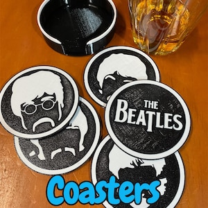 May include: A set of black and white coasters featuring images of The Beatles band members and the band's logo. The coasters are round and sit on a wooden surface, with a black coaster holder. The word "Coasters" is written in blue.