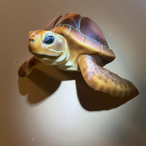 May include: A detailed, three-dimensional turtle sculpture. The turtle features a brown shell with yellow and orange accents, a yellow head, and flippers. The sculpture is mounted on a neutral-colored surface, showcasing the turtle's realistic details and textures.