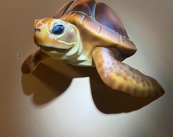 Può includere: Una scultura dettagliata di una tartaruga tridimensionale. La tartaruga presenta un guscio marrone con dettagli gialli e arancioni, una testa gialla e pinne. La scultura è montata su una superficie di colore neutro, che mostra i dettagli e le trame realistiche della tartaruga.