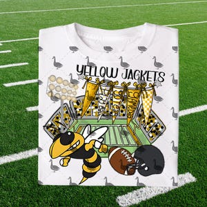 Può includere: T-shirt bianca con un design grafico raffigurante un'ape dei cartoni animati gialla e nera, un pallone da football e un casco nero. La maglietta riporta le scritte "Yellow Jackets" e "Gameday". Il design include un campo da football e uno stadio.