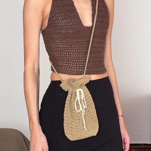 May include: A dark brown crochet halter top with a deep V-neckline paired with a tan crochet drawstring pouch bag featuring a white cord closure and a long thin strap worn across the body over a plain black mini skirt.